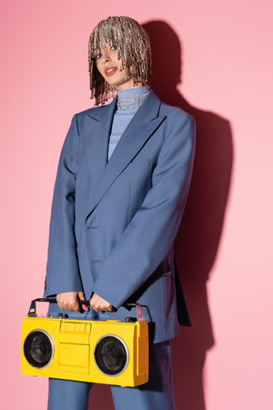 Fashionable woman in luxury headwear holding boombox and looking at camera on pink background