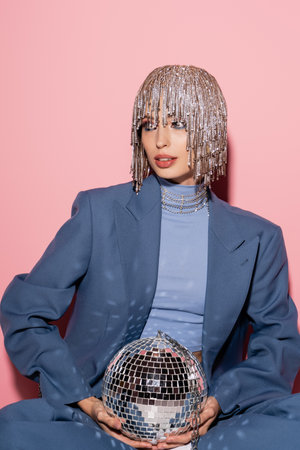 Fashionable young woman in blue suit and jewelry headwear holding disco ball on pink