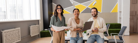 cheerful multicultural business colleagues with documents and gadgets looking at camera in modern...