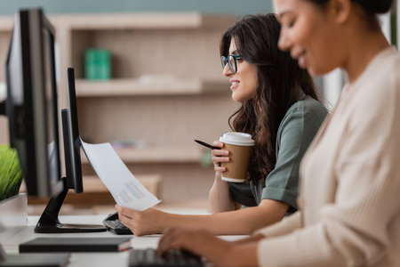 side view of manager in eyeglasses holding paper cup and document near multiracial woman working on...