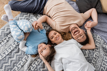 top view of excited parents and cheerful kid lying on bed at homeの写真素材
