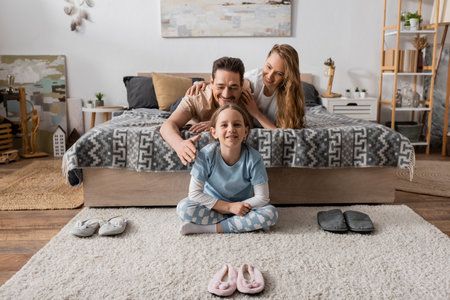 positive child sitting on carpet around slippers near happy parents on bedの写真素材