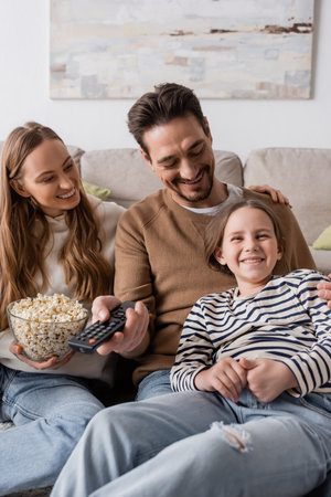 happy man holding remote controller while looking at daughter near wife with popcornの写真素材