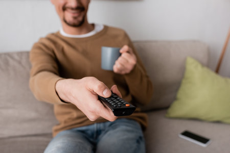 cropped view of cheerful man holding remote controller and cup of coffee in living room