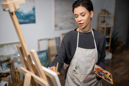 Short haired artist in apron holding palette while painting on blurred canvasの写真素材