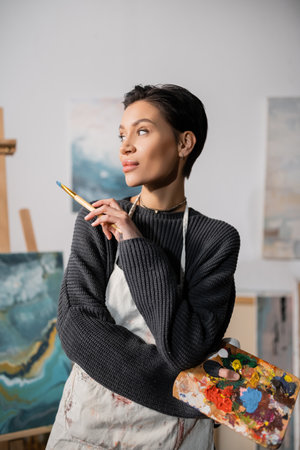 Short haired artist in sweater and apron holding palette with paints and paintbrushの写真素材