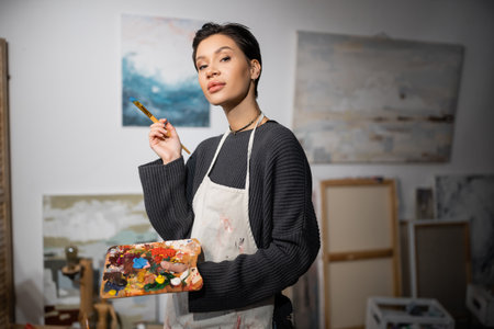 Brunette artist looking at camera while holding paintbrush and palette in blurred workshopの写真素材
