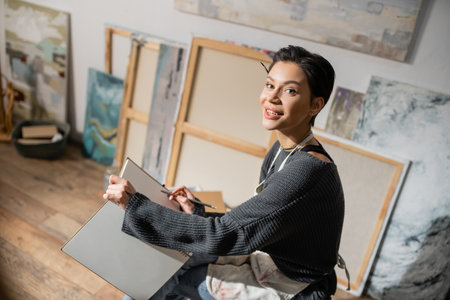 Young short haired artist holding pencil and sketchbook in studio