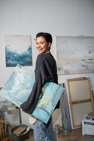 Happy artist looking at camera and holding painting in studio