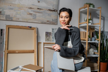Young artist holding pencil and sketchbook and looking at camera in workshop