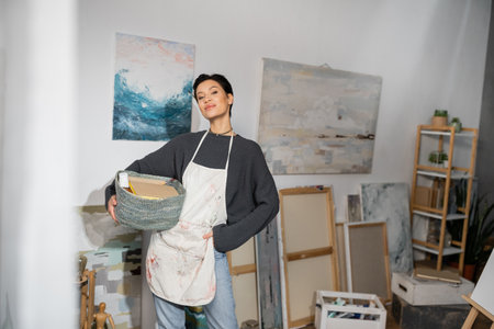 Short haired artist in apron holding basket with equipment near drawings in studioの写真素材