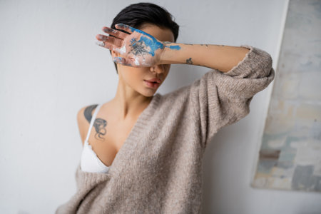Sensual artist in bra and sweater holding hand in paint near face in workshopの写真素材