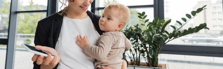 cheerful businesswoman holding toddler girl and cellphone with blank screen in office, banner