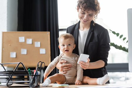 little baby in romper sitting on desk near smiling mom in black blazer using mobile phone in officeの写真素材