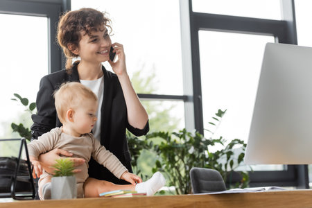 toddler girl sitting on office desk near happy mother talking on smartphone in officeの写真素材