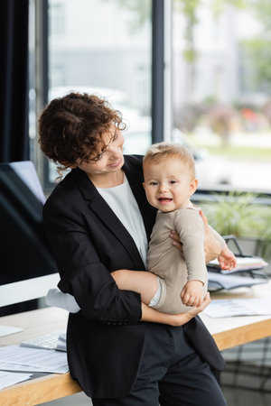 smiling businesswoman standing with little daughter near workplace in officeの写真素材