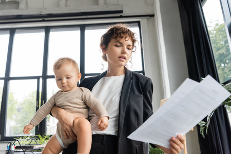 office manager in black blazer looking at papers while holding toddler baby in officeの写真素材