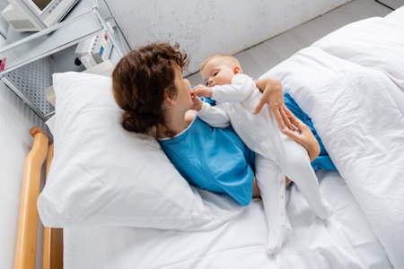high angle view of baby in romper touching face of mother on bed in clinicの写真素材