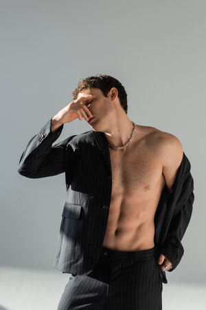 shirtless man in black blazer and pants covering face with hand while standing on grey background