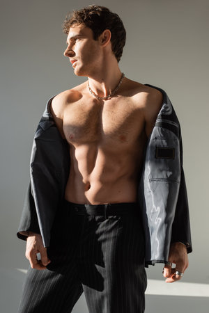 sexy and muscular man looking away while posing in pants and black blazer on shirtless body on grey backgroundの写真素材