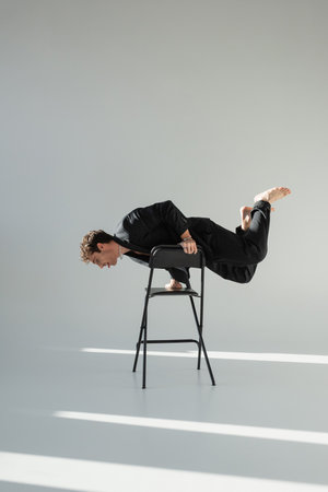 full length of barefoot man in black suit doing trick on chair on grey background
