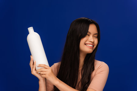 Cheerful asian woman holding bottle of shampoo isolated on blue