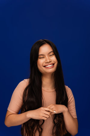 Pleased young asian woman in beige t-shirt standing isolated on blue