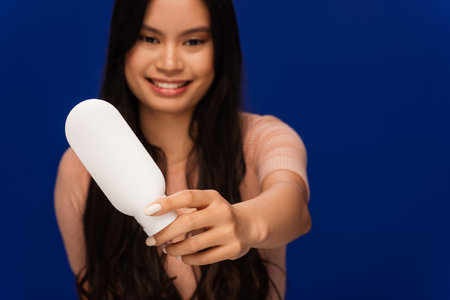 Blurred asian woman showing cosmetic lotion isolated on blue