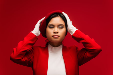 Fashionable asian model in beret touching head isolated on redの写真素材
