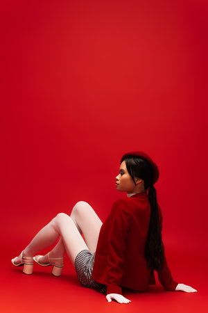 Side view of stylish asian model in white tights and gloves looking away while sitting on red backgroundの写真素材