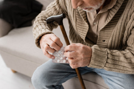 cropped view of senior man with parkinson disease sitting with walking cane and pills on couch at homeの写真素材
