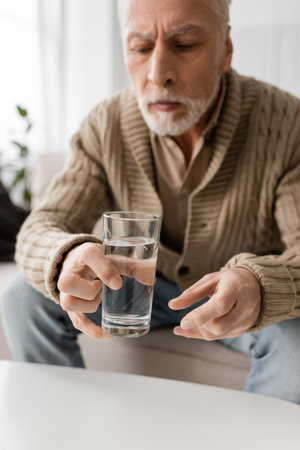 blurred man with parkinson syndrome and tremor in hands sitting and holding glass of water at homeの写真素材