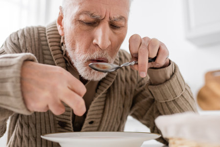 aged man suffering from parkinsonian syndrome holding spoon in trembling hand while having dinner at homeの写真素材