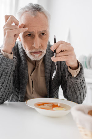 depressed man with parkinsonian syndrome looking away while holding spoon near soup in kitchenの写真素材