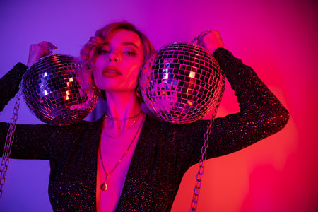 young woman with blonde hair standing in black dress and holding chains with disco balls on purple and pinkの写真素材