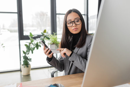 Brunette asian interior designer looking at computer and pointing at smartphone in studioの写真素材