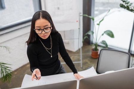 Asian designer pointing at computer near blueprint on table in studioの写真素材