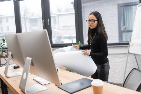 Asian interior designer holding blueprint near computers in officeの写真素材
