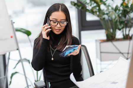 Smiling asian designer talking on smartphone and holding color samples near blueprint in office