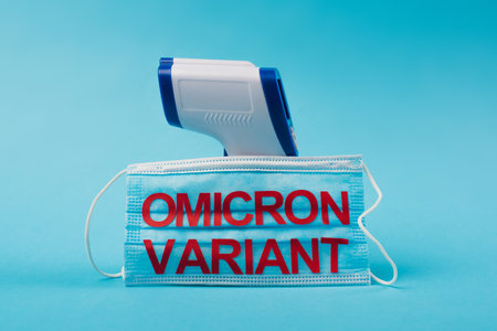 Medical mask with omicron variant lettering near pyrometer on blue backgroundの写真素材