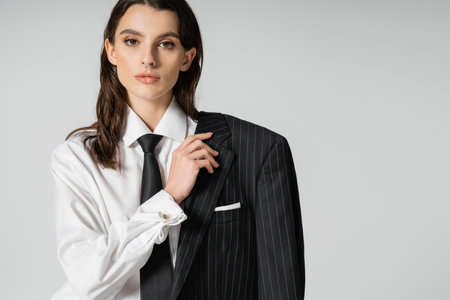 trendy woman in white shirt and tie holding black striped blazer and looking at camera isolated on...