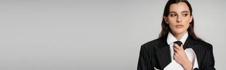pretty woman in fashionable oversize attire adjusting tie and looking away isolated on grey, banner