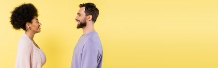 side view of curly african american woman and bearded man smiling at each other isolated on yellow,...