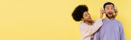 curly african american woman putting wireless headphones on amazed man isolated on yellow, bannerの写真素材