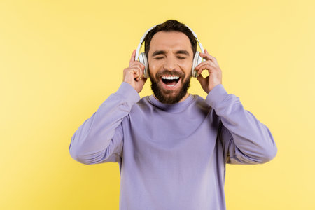 excited bearded man adjusting wireless headphones and laughing with closed eyes isolated on yellowの写真素材
