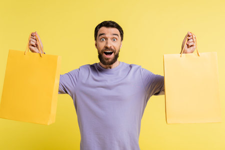 amazed bearded man in purple pullover holding shopping bags and looking at camera isolated on yellow