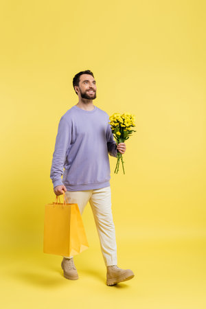 full length of happy and stylish man walking with shopping bags and flowers on yellow backgroundの写真素材