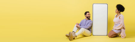 full length of cheerful interracial couple sitting near huge template of smartphone on yellow background, bannerの写真素材