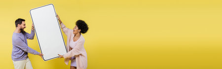 astonished interracial couple holding big template of smartphone isolated on yellow, bannerの写真素材