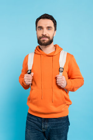 bearded brunette student in orange hoodie holding backpack and looking at camera isolated on blueの写真素材